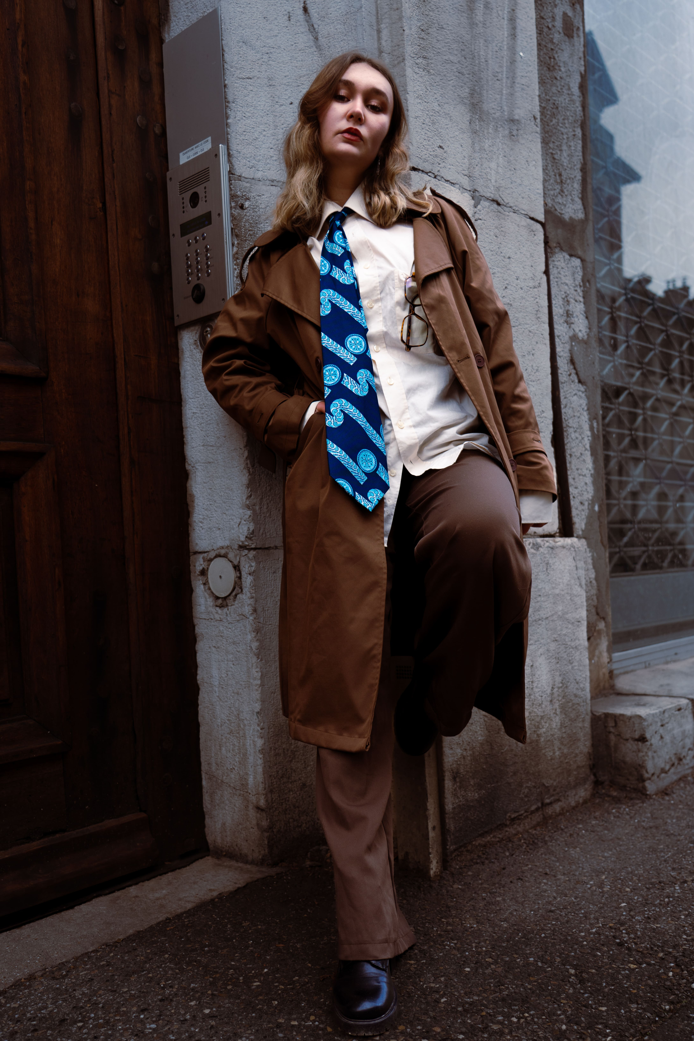 photo de mode magazine femme adoosée à un mur une jambe relevée en costume et veste marron avec une chemise blanche et une cravate bleue. La femme est jeune est b;londe photographge de mode chambéry grenoble