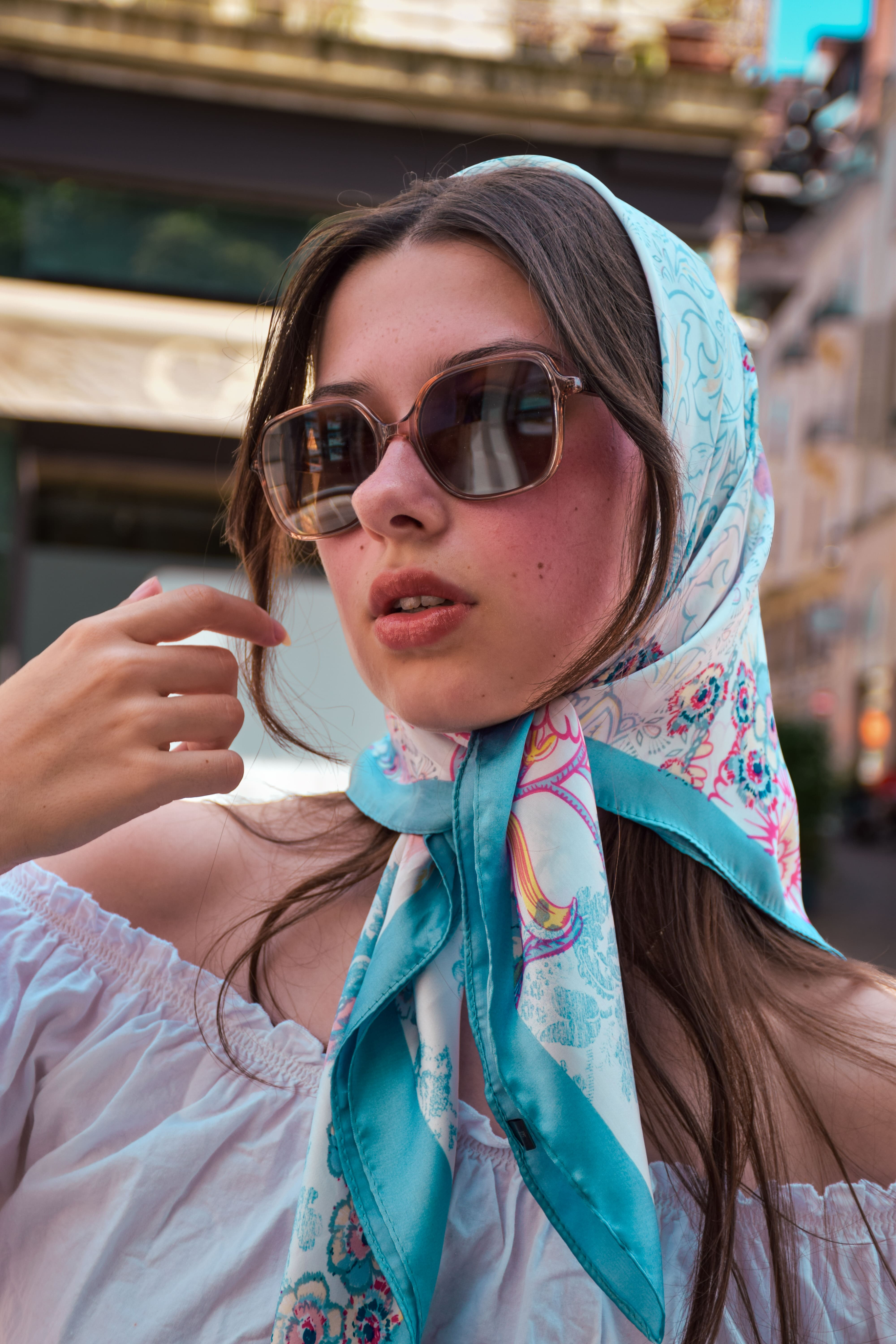 photo de mode femme devant un immeuble prise en portrait de près on voit ses épaules et le haut de sa tête. Elle a les cheveux noirs et un foulard sur la tête style années 60 et des lunettes de soleil. photographge de mode chambéry grenoble