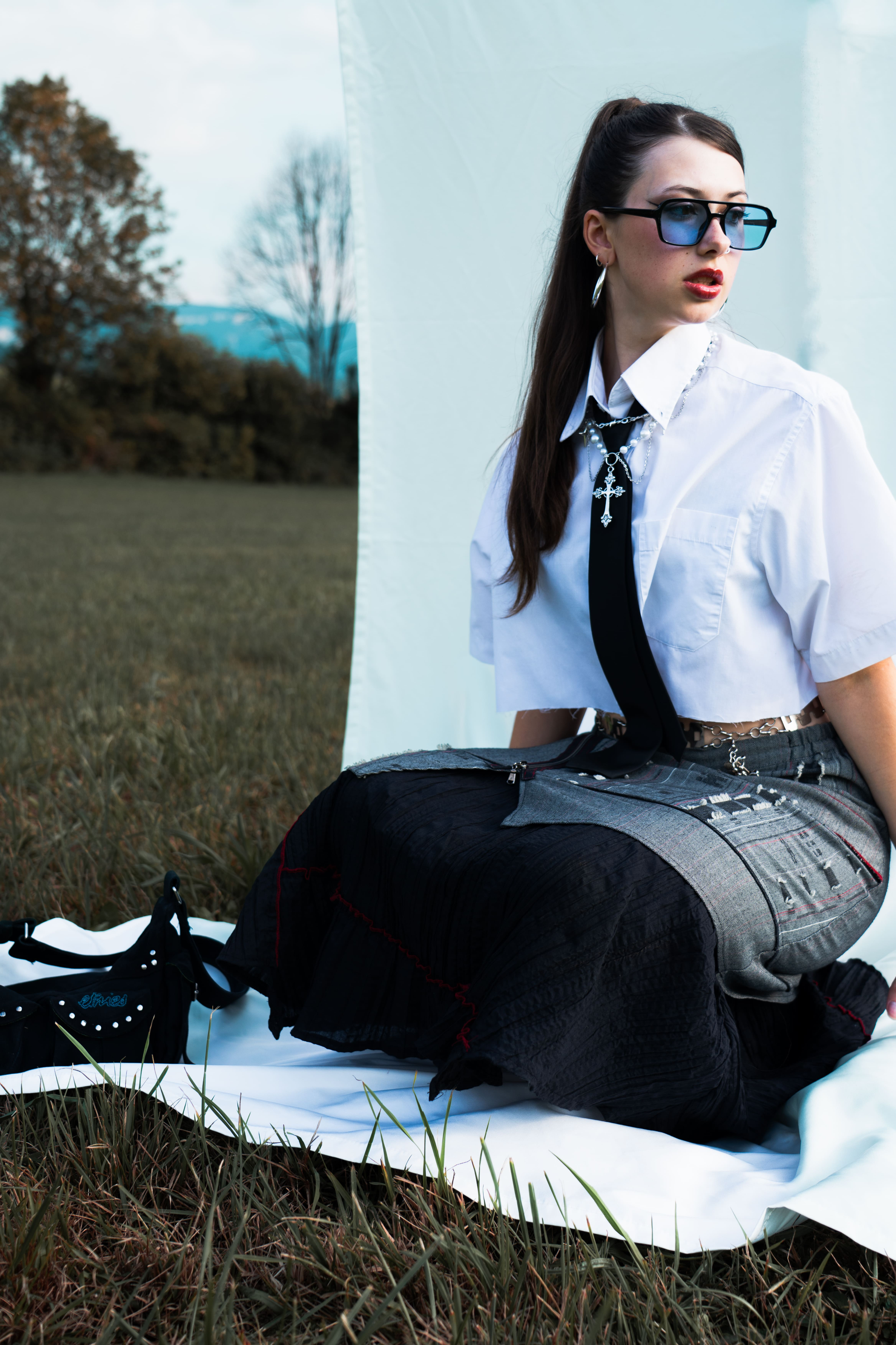 femme accroupie dans un champs sur un drap blanc en jupe chemise blanche cravate avec lunettes de soleil et des cheveux longs noirs en queue de cheval avec son sac a main a coté d'elle photographge de mode chambéry grenoble