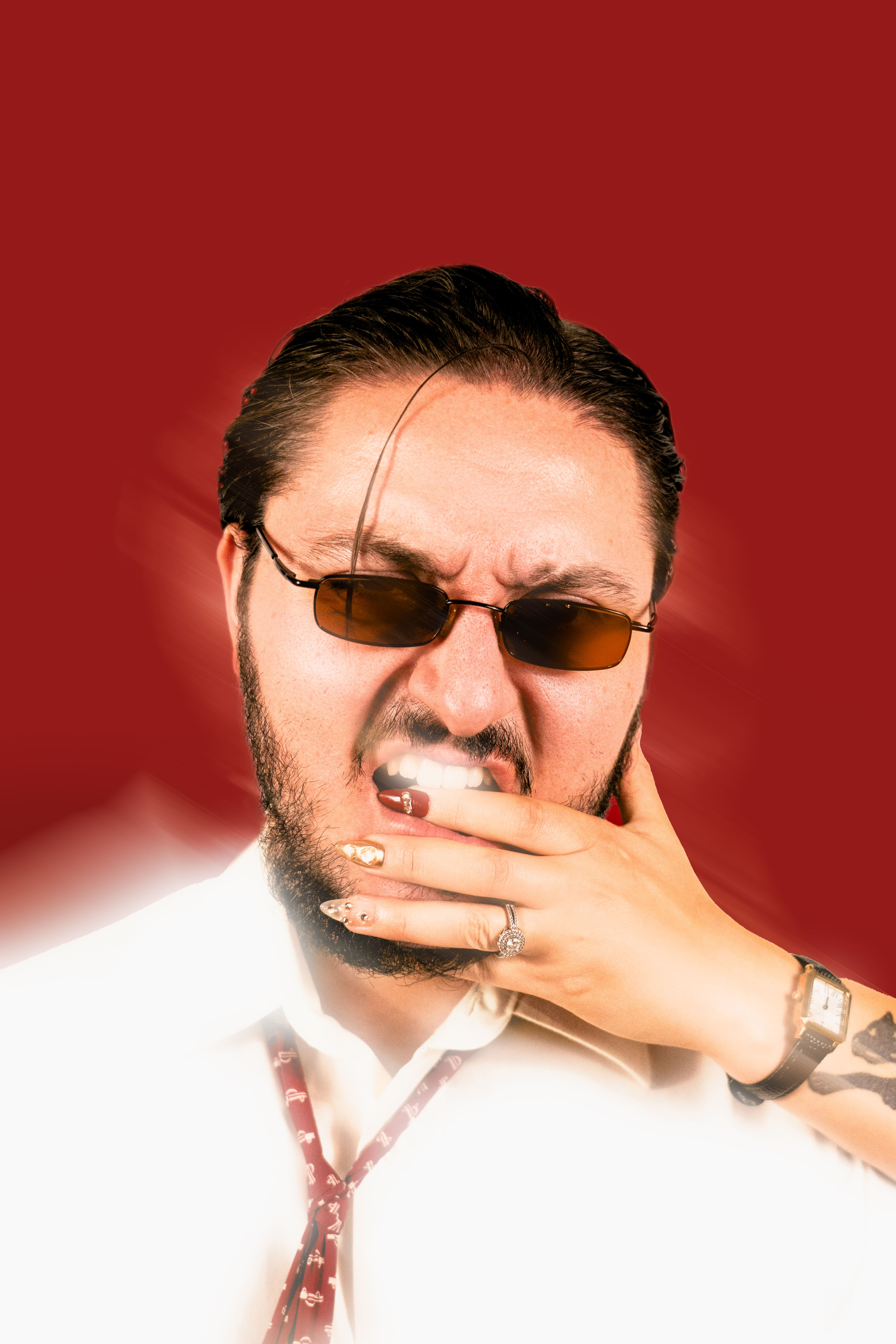 photo homme en chemise blanche et cravate rouge devant un fond rouge. Portrait de près, il a des lunettes de soleil les cheveux gominés marrons et une barbe et il mord une main de femme avec des ongles rouges. une création originale pauline artbook photographe chambéry et grenoble