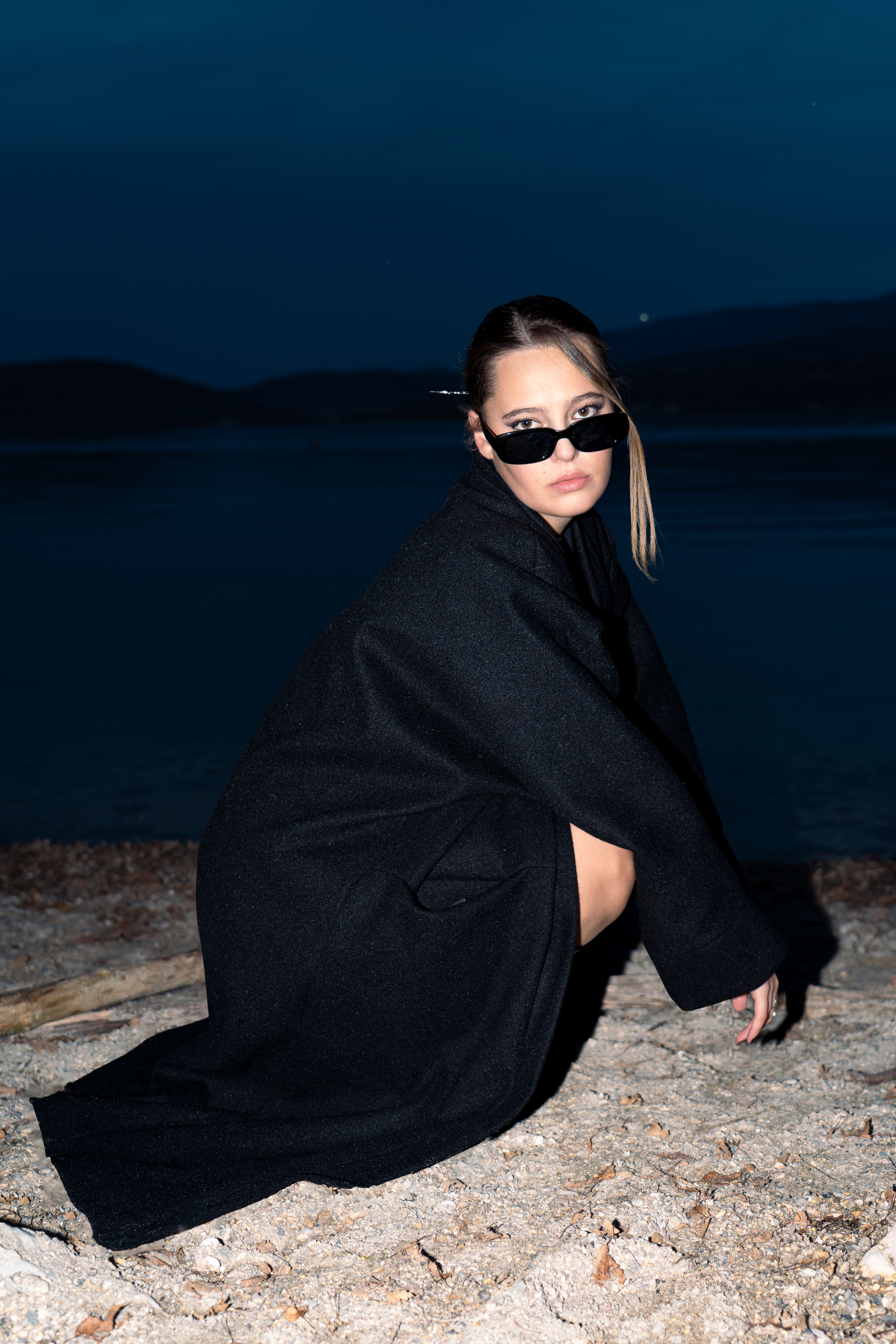 femme accroupie devant un lac . style mode femme fatale long manteau noir avec lunettes de soleil noires femme blonde. photographge de mode chambéry grenoble